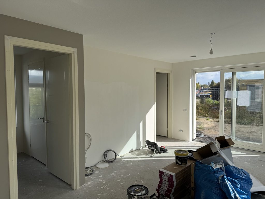 Nieuwbouwwoonkamer tijdens de afwerking, met verfemmers en dozen op de vloer en een schuifpui naar de tuin